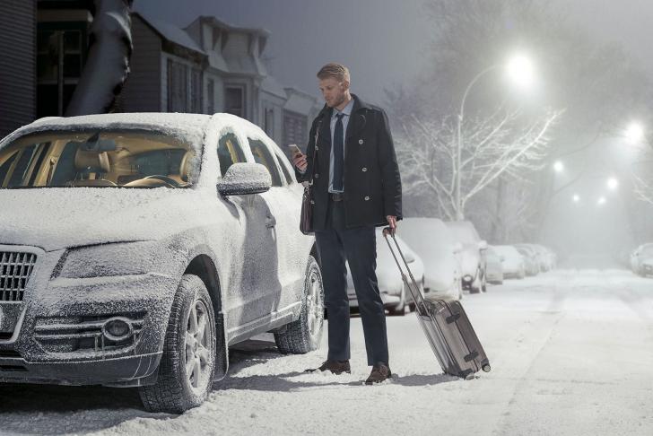 Businessmann steht mit Smartphone am eingeschneiten Auto, nur die Scheiben sind abgetaut